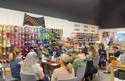 Group of people in a yarn store with shelves of colorful yarn and knitting supplies.